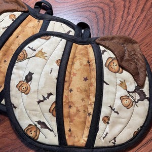 Peut inclure: Deux maniques en tissu matelassé en forme de citrouille avec une bordure noire et une feuille en velours côtelé marron. Le tissu présente un fond crème et beige avec un motif répétitif de citrouilles d'Halloween, chauves-souris et sorcières.