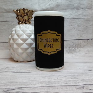 May include: A black wipe dispenser with a gold label that reads "DISINFECTING WIPES". The dispenser is cylindrical with a white top. A white and gold pineapple-shaped container is to the left of the dispenser.