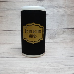 May include: A cylindrical container for disinfecting wipes. The container is black with a gold label that reads "DISINFECTING WIPES". The top and bottom of the container are white. The container is on a light wood-grain surface.