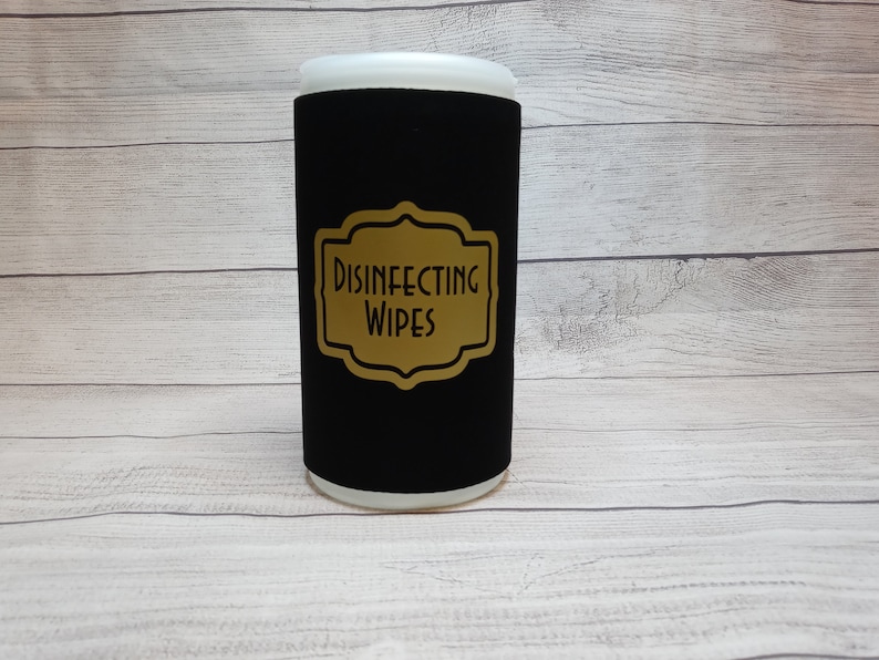 May include: A cylindrical container for disinfecting wipes. The container is black with a gold label that reads "DISINFECTING WIPES". The top and bottom of the container are white. The container is on a light wood-grain surface.