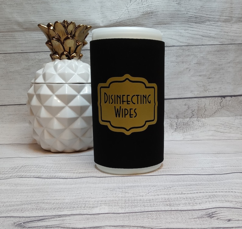 May include: A black wipe dispenser with a gold label that reads "DISINFECTING WIPES". The dispenser is cylindrical with a white top. A white and gold pineapple-shaped container is to the left of the dispenser.