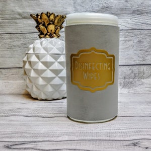 May include: A gray cylindrical container with a white lid and a gold label that reads "DISINFECTING WIPES." A white pineapple-shaped decorative jar with a gold top is to the left. The items are on a light wood-grain surface.