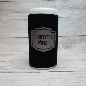 May include: A black cylindrical container with a white top, labeled "DISINFECTING WIPES" in a silver, decorative frame. The container is designed to hold disinfecting wipes.