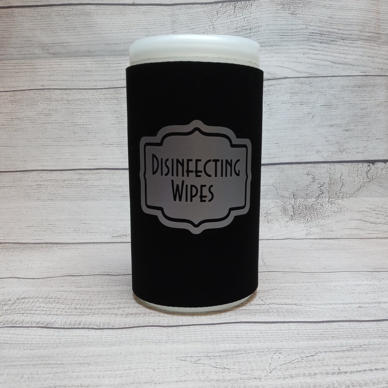 May include: A black cylindrical container with a white top, labeled "DISINFECTING WIPES" in a silver, decorative frame. The container is designed to hold disinfecting wipes.