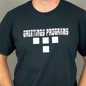 May include: A dark navy blue short-sleeved crew neck t-shirt featuring the white text GREETINGS PROGRAMS printed in a bold, angular font. Below the text are four white square shapes arranged in a T-like pattern on the chest area.