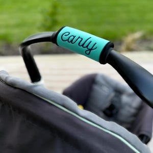 May include: A bright turquoise handle cover for a stroller, featuring the name Carly embroidered in a dark script font. The cover is wrapped around a black curved stroller handle, with a gray fabric seat visible in the blurred background.