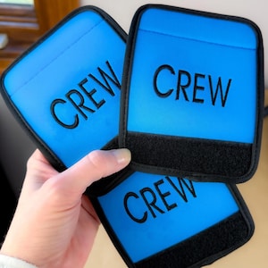 May include: Three blue rectangular patches with the word "CREW" embroidered in black. Each patch has a black border and a black Velcro strip at the bottom. The patches are held in a person's hand.