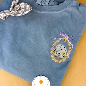 May include: A blue t-shirt with an embroidered design of a gold-framed globe with a purple bow. A patterned bow tie is visible in the upper left corner. The shirt has a small tag with the size L-G.