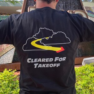 May include: Black t-shirt with a graphic of clouds, a yellow path, and a red arrow. The text "CLEARED FOR TAKEOFF" is printed below the graphic in white and gray. The shirt is made of a soft material.