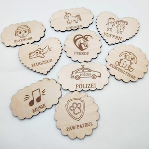 May include: A collection of wooden cutouts with various engraved designs. The shapes include ovals and hearts, featuring images of a unicorn, a plane, a police car, and a musical note. Text includes "PLAYMOBIL", "EINHORN", "PUPPEN", "FLUGZEUG", "PFERDE", "KUSCHELTIERE", "POLIZEI", "MUSIK", and "PAW PATROL".