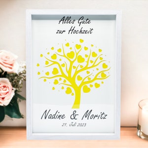 May include: White framed art print with a yellow tree design filled with heart-shaped leaves. The text "Alles Gute zur Hochzeit" is at the top, with names and a date at the bottom. A wedding-themed gift.