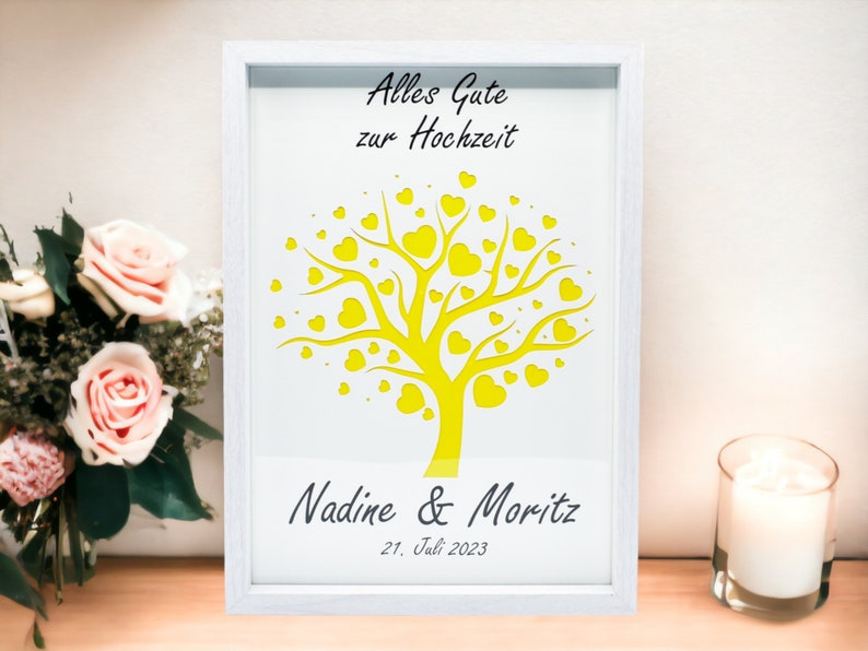 May include: White framed art print with a yellow tree design filled with heart-shaped leaves. The text "Alles Gute zur Hochzeit" is at the top, with names and a date at the bottom. A wedding-themed gift.