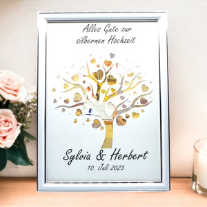 May include: Framed artwork with a silver frame, featuring a tree design with heart-shaped leaves. The text "Alles Gute zur silbernen Hochzeit" is at the top, with names "Sylvia & Herbert" and the date "10. Juli 2023" at the bottom.