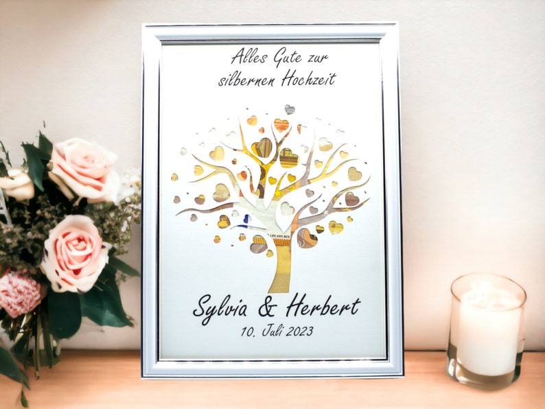 May include: Framed artwork with a silver frame, featuring a tree design with heart-shaped leaves. The text "Alles Gute zur silbernen Hochzeit" is at the top, with names "Sylvia & Herbert" and the date "10. Juli 2023" at the bottom.