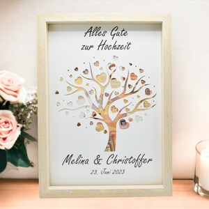 May include: A framed artwork with a tree design, featuring heart-shaped cutouts and currency details. The text "Alles Gute zur Hochzeit" is at the top, with names and a date below. The frame is light-colored, and the artwork is displayed on a wooden surface.