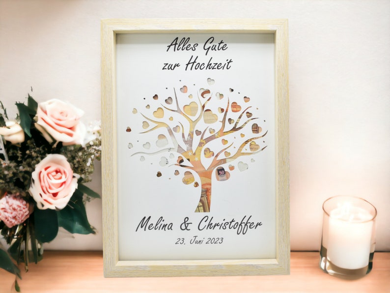 May include: A framed artwork with a tree design, featuring heart-shaped cutouts and currency details. The text "Alles Gute zur Hochzeit" is at the top, with names and a date below. The frame is light-colored, and the artwork is displayed on a wooden surface.