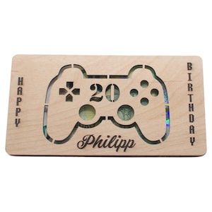 May include: A light-colored wooden birthday card with a cutout of a video game controller. The number "20" is in the center, with the name "Philipp" below. The words "HAPPY" and "BIRTHDAY" are vertically aligned on the sides.