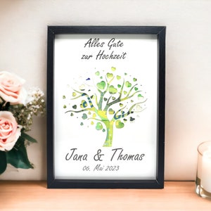 May include: A framed print with a black border features a tree design with green heart-shaped leaves. The text "Alles Gute zur Hochzeit" is at the top, with "Jana & Thomas" and the date "06. Mai 2023" at the bottom. A bouquet of pink roses and a lit candle are in the background.