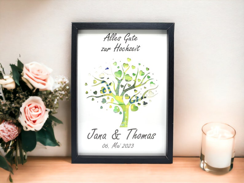 May include: A framed print with a black border features a tree design with green heart-shaped leaves. The text "Alles Gute zur Hochzeit" is at the top, with "Jana & Thomas" and the date "06. Mai 2023" at the bottom. A bouquet of pink roses and a lit candle are in the background.