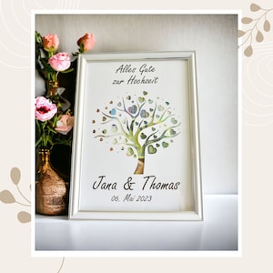 May include: A framed artwork with a tree design, featuring heart-shaped leaves and a trunk made of currency. The text "Alles Gute zur Hochzeit" is at the top, with "Jana & Thomas" and the date "06. Mai 2023" below. The frame is silver.