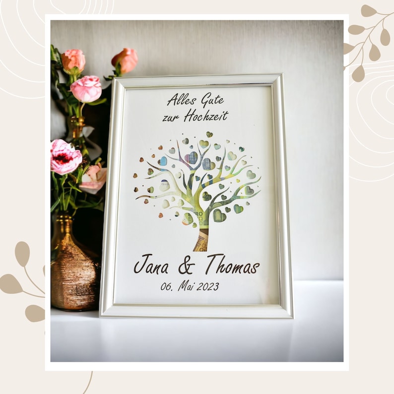 May include: A framed artwork with a tree design, featuring heart-shaped leaves and a trunk made of currency. The text "Alles Gute zur Hochzeit" is at the top, with "Jana & Thomas" and the date "06. Mai 2023" below. The frame is silver.