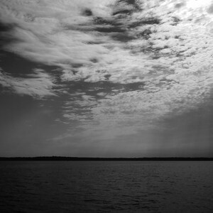 Op de afbeelding: Een zwart-wit landschapsfoto met een watermassa onder een bewolkte hemel. De lucht is gevuld met getextureerde wolken en het water weerspiegelt de donkere tinten van de lucht. De horizon is zichtbaar.