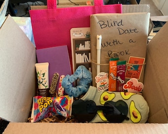 May include: A brown cardboard box filled with a variety of items for a "Blind Date with a Book" themed gift. The box includes a wrapped book, a pink tote bag, a purple notebook, a tube of hand cream, a pack of ring pops, a pair of sunglasses, a pair of avocado socks, a blue scrunchie, a pack of nail stickers, a pack of book-themed stickers, two packets of Crystal Light, and a "My Weekend is All Booked" sticker.