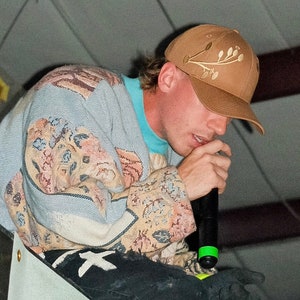 Peut inclure: Une personne portant une casquette de baseball marron avec un motif floral, une veste bleu clair et beige à motifs, un t-shirt turquoise et un jean noir avec des graphiques blancs. La personne tient un microphone.