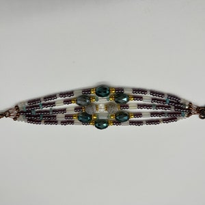 May include: A multi-strand bracelet with dark purple, white, and light blue beads. The center of the bracelet features a cluster of dark blue faceted beads and a gray stone.