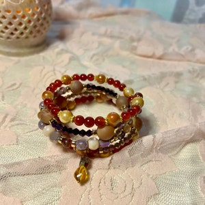 May include: A multi-strand bracelet with red, orange, brown, purple, and gold beads. The bracelet is wrapped around itself and has a gold charm hanging from the bottom.
