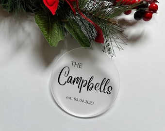 May include: A round, frosted acrylic ornament featuring the black printed text THE Campbells and est. 05.04.2023. The ornament is suspended by a red ribbon and is positioned beneath green pine needles and red berries.
