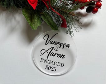 May include: A round, frosted acrylic ornament featuring the black printed text Vanessa & Aaron ENGAGED 2025. The ornament is suspended by a red ribbon and is positioned beneath green pine needles and red berries.