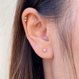 May include: A gold-toned ear piercing set featuring a small pink stud earring in the lobe, a gold hoop with a clear stone in the rook, and two small clear stud earrings positioned along the upper cartilage edge of the ear.