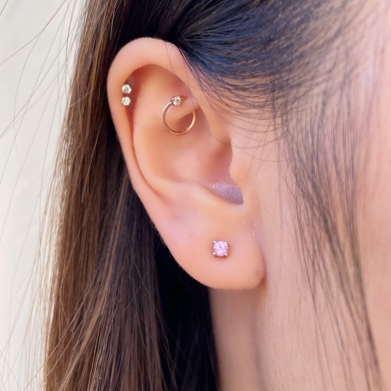 May include: A gold-toned ear piercing set featuring a small pink stud earring in the lobe, a gold hoop with a clear stone in the rook, and two small clear stud earrings positioned along the upper cartilage edge of the ear.