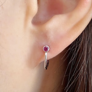 Puede incluir: Primer plano de un pendiente de aro plateado con una pequeña gema roja y redonda. El pendiente está sujeto a una oreja. La gema está engastada en un bisel plateado.