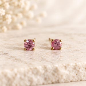 May include: A pair of stud earrings featuring round, faceted pink gemstones set in gold metal. Each stone is secured by four small prongs, creating a classic design that highlights the vibrant pink colour and the reflective quality of the cut stones.