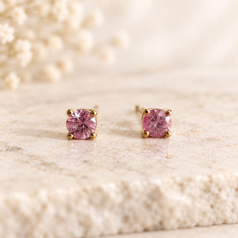 May include: A pair of stud earrings featuring round, faceted pink gemstones set in gold metal. Each stone is secured by four small prongs, creating a classic design that highlights the vibrant pink color and the reflective quality of the cut stones.