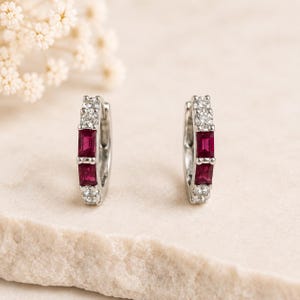 May include: A pair of silver hoop earrings featuring two rectangular red rubies set vertically in the center, flanked by two clear round stones at the top and bottom of each hoop, creating a symmetrical design with a polished metallic finish.