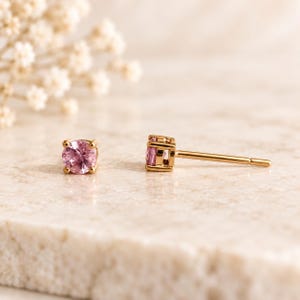 May include: A pair of stud earrings featuring round, faceted pink gemstones set in gold metal prongs. One earring is shown from the front, displaying the circular pink stone, while the other is positioned to the side, revealing the gold post.