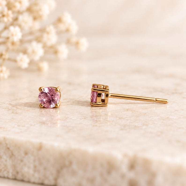 May include: A pair of stud earrings featuring round, faceted pink gemstones set in gold metal prongs. One earring is shown from the front, displaying the circular pink stone, while the other is positioned to the side, revealing the gold post.