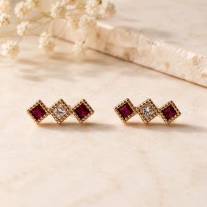 May include: A pair of gold stud earrings, each featuring three square-cut stones arranged in a horizontal row. The outer stones are deep red, while the center stone is clear and colorless. Each stone is set within a beaded gold metal frame.