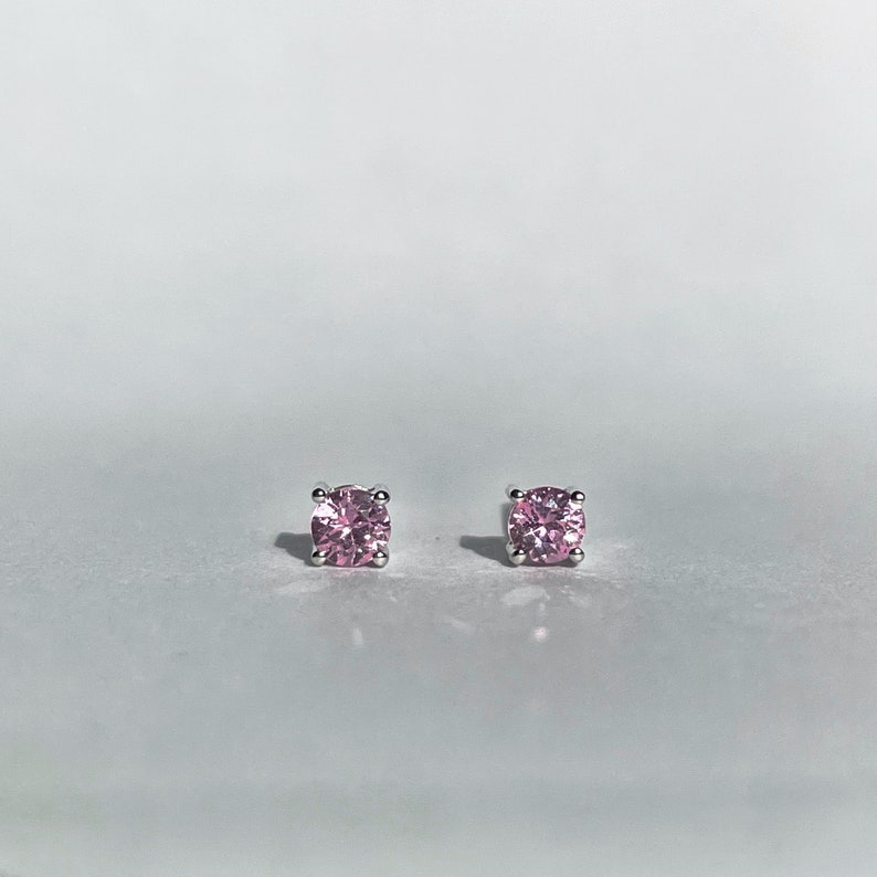 May include: A pair of stud earrings featuring round, faceted pink gemstones set in four-prong silver-toned metal settings. The stones exhibit a brilliant, reflective quality with a clear, vibrant pink hue, securely held by the metallic prongs.