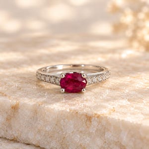 May include: A silver-toned ring featuring a central oval-cut deep red ruby held by four prongs. The band is accented with a row of small, round, clear pavé-set stones extending along both sides of the central gemstone setting.