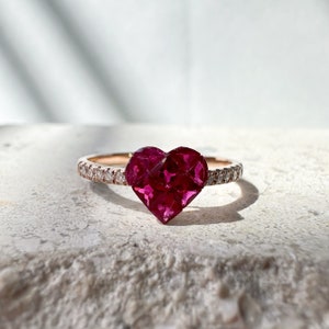 Op de afbeelding: Een roségouden ring met een grote, facetgeslepen, hartvormige robijn. De band is bezet met kleine, heldere edelstenen. De ring ligt op een getextureerd, lichtgekleurd oppervlak met schaduwen.