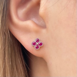May include: A pair of small, silver stud earrings with a four-leaf clover design. Each leaf is set with a round, red ruby.