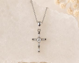 May include: A silver-toned metal cross pendant necklace featuring a central round brilliant-cut clear stone surrounded by four baguette-cut clear stones arranged in a cross shape, suspended from a delicate matching silver-toned metal chain.