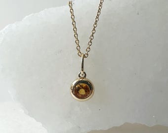 Op de afbeelding: Een gouden ketting met een klein rond hanger met een gele edelsteen.