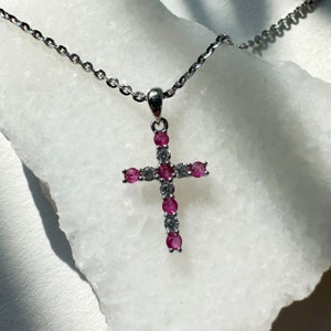 Può includere: Una collana con catena d'argento con un piccolo ciondolo a forma di croce. La croce è decorata con pietre preziose rosa e bianche.