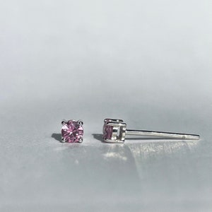 May include: A pair of stud earrings featuring round, faceted pink stones set in a four-prong silver-toned metal setting. One earring is shown from the front, while the other is positioned on its side to display the post and the basket setting.