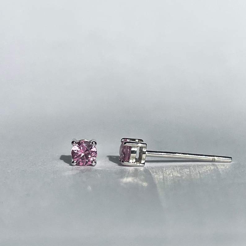 May include: A pair of stud earrings featuring round, faceted pink stones set in a four-prong silver-toned metal setting. One earring is shown from the front, while the other is positioned on its side to display the post and the basket setting.
